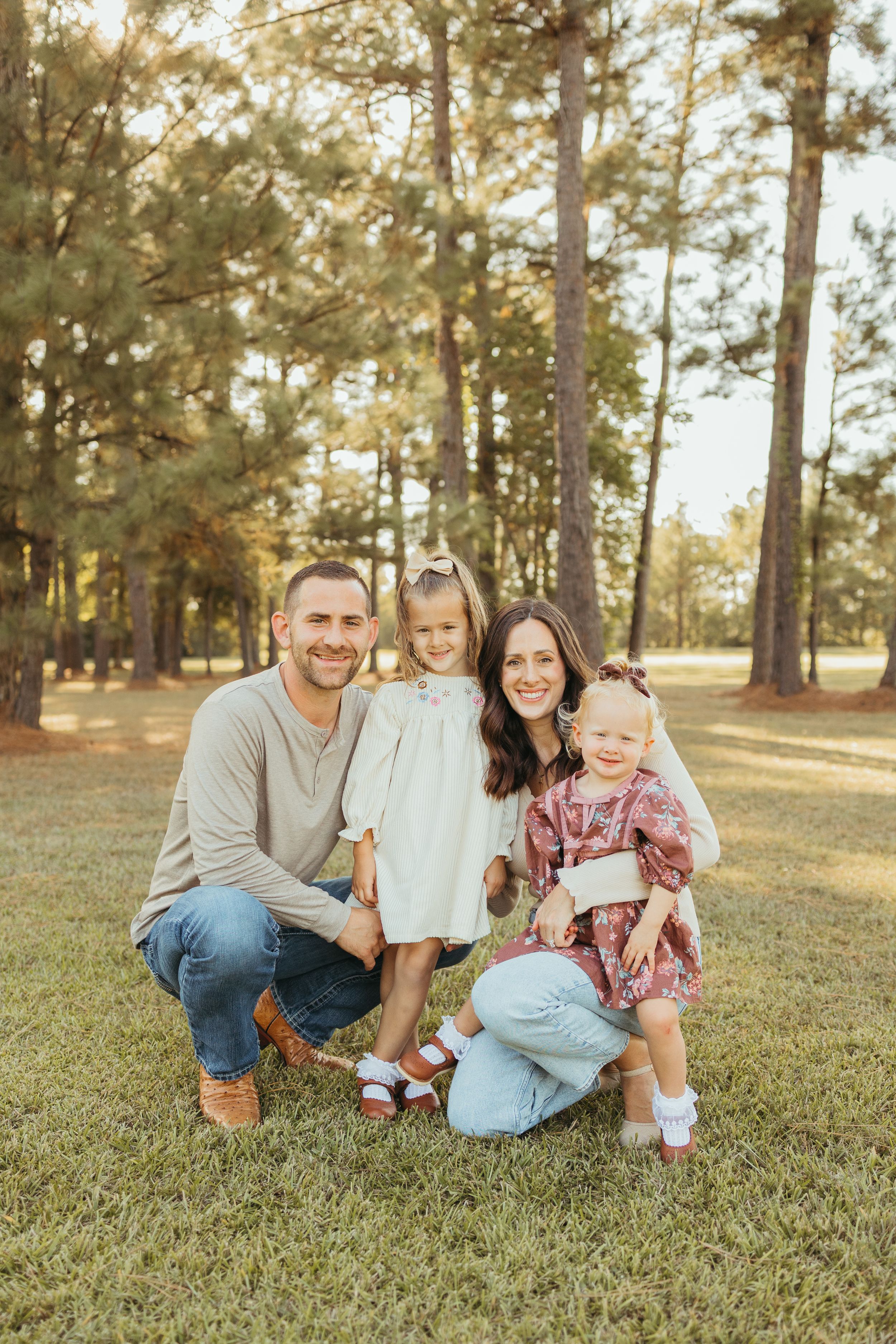 Family of four poses smiling in a sunny pine forest park, parents crouching with two young daughters in coordinated outfits.