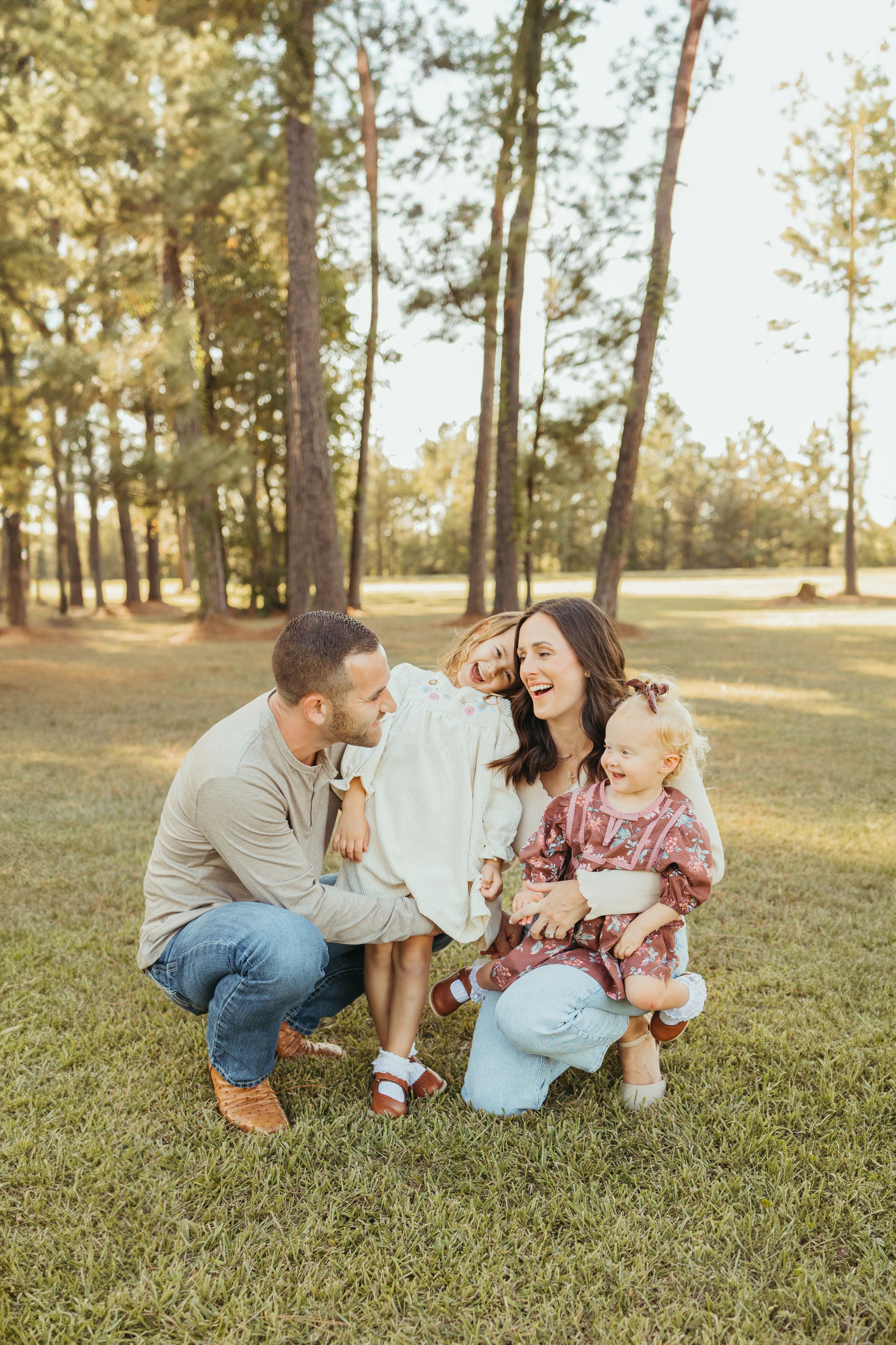 Happy family of four laughing together outdoors in a sunlit pine forest, parents crouching with two little girls.