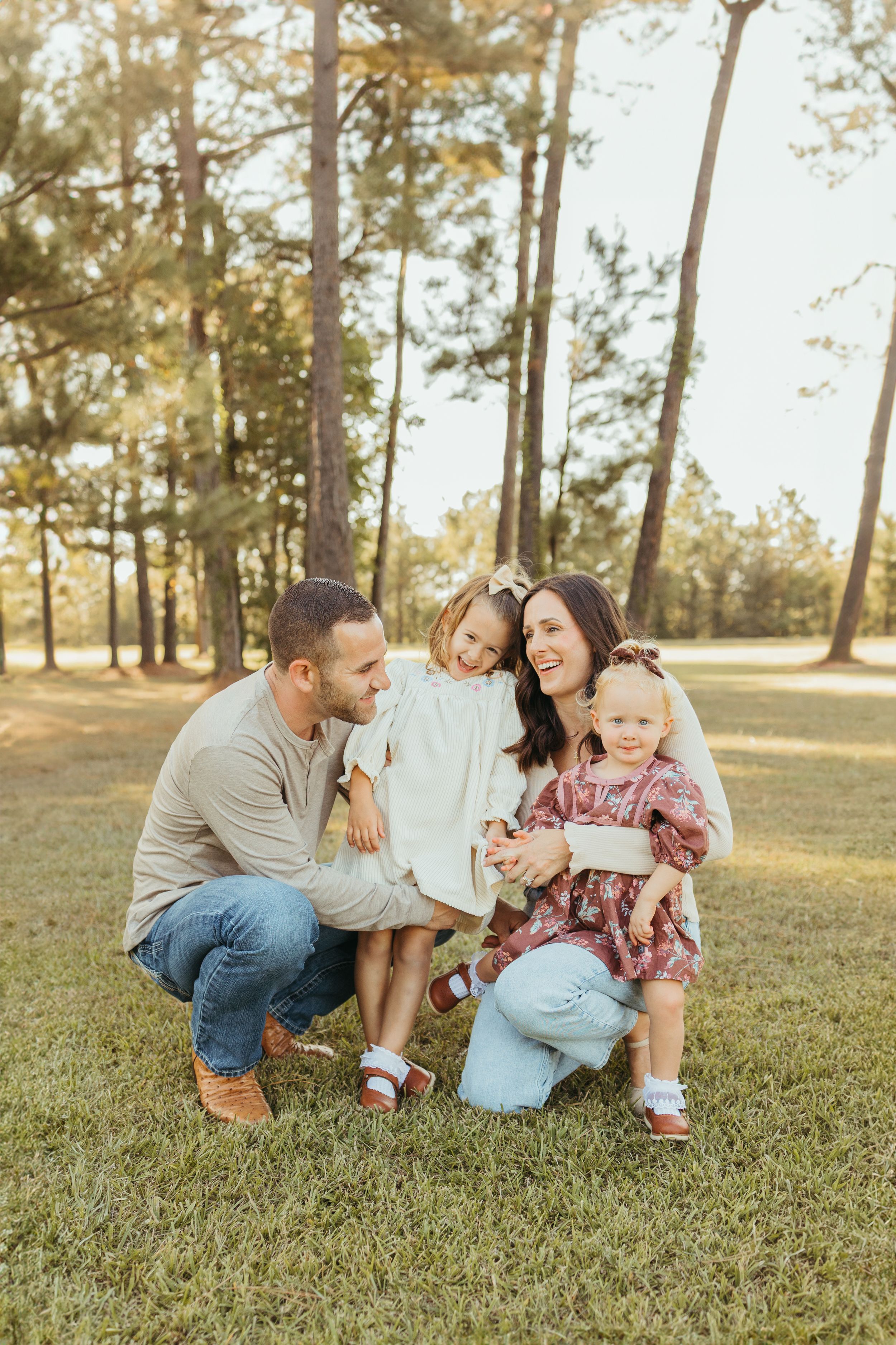 Happy family crouching together on grass, parents smiling at two little girls in a sunlit pine forest.