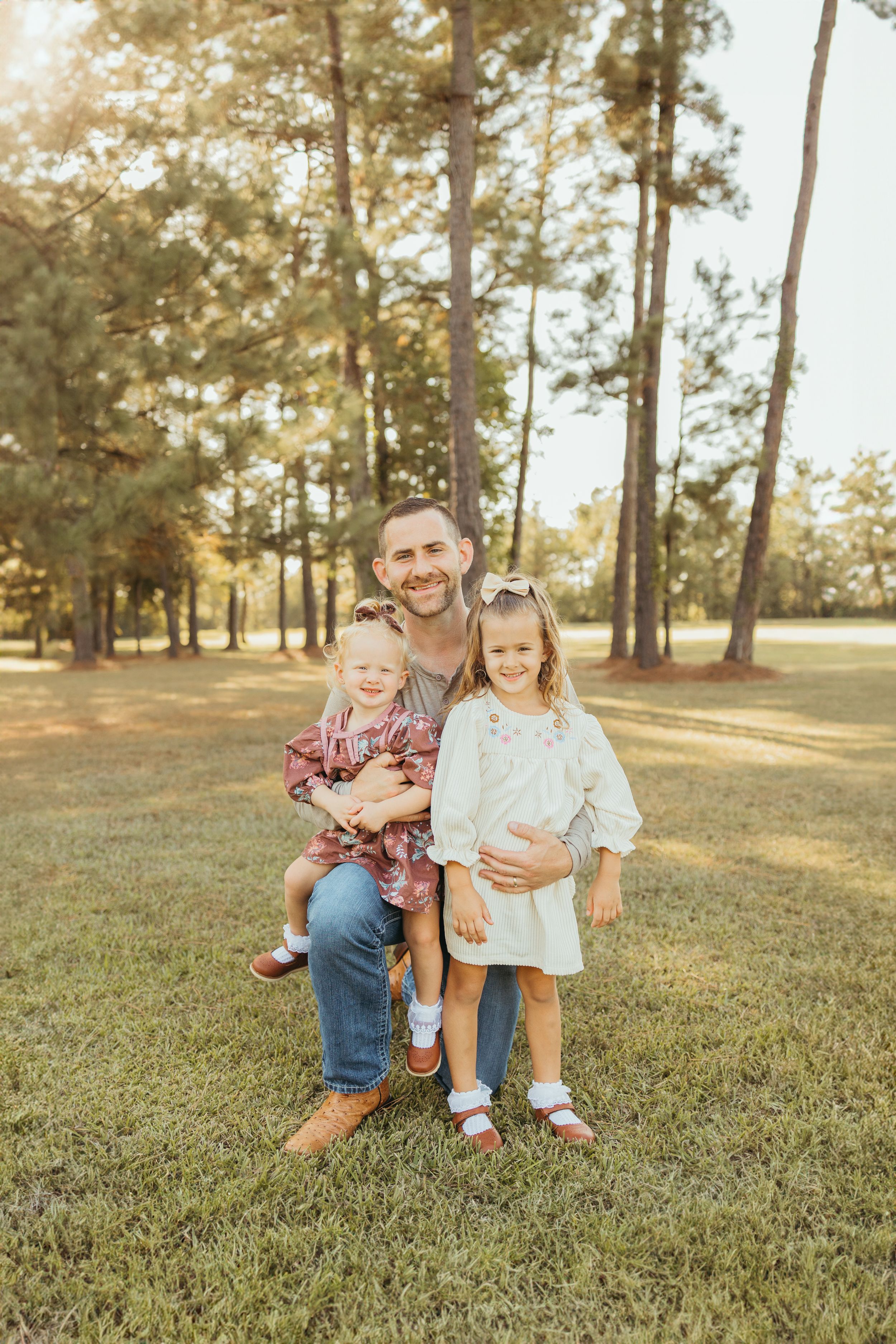 A smiling father poses outdoors with two young daughters in a sunny park surrounded by tall pine trees.