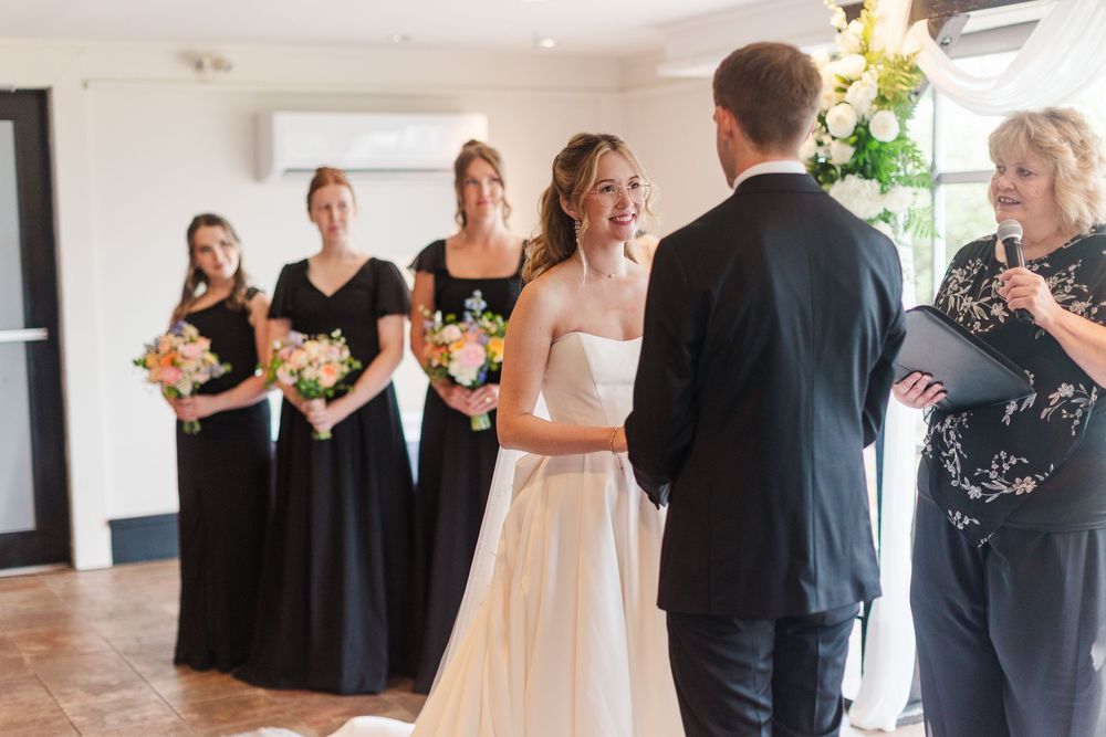 Bride and groom exchange vows at indoor wedding ceremony with bridesmaids in black dresses holding pink bouquets.