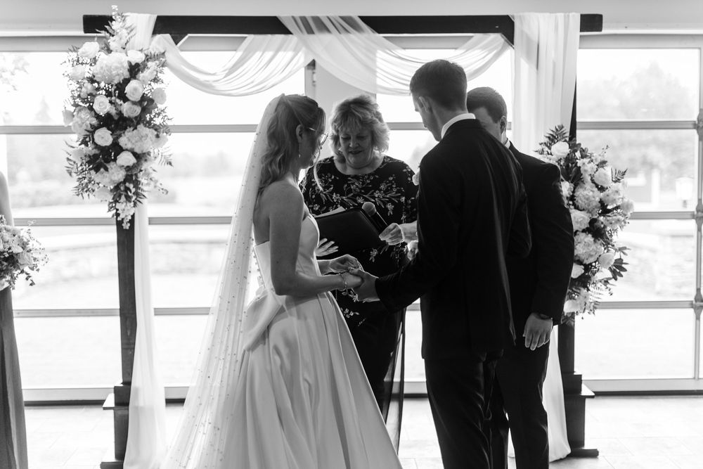 Black and white photo of a bride and groom exchanging vows with an officiant at a floral-decorated wedding altar.