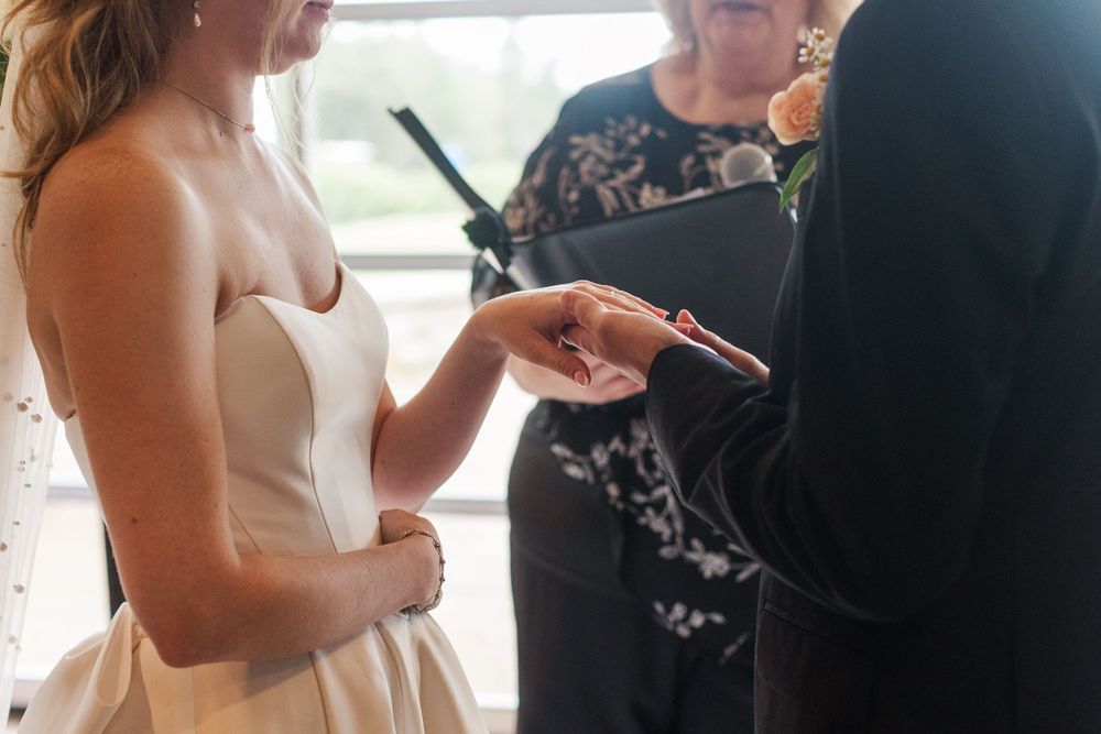 Groom placing a wedding ring on bride's finger during indoor ceremony, officiant in background.
