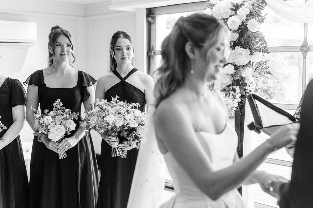 Black and white photo of bride adjusting dress while two bridesmaids in dark gowns hold bouquets at wedding.