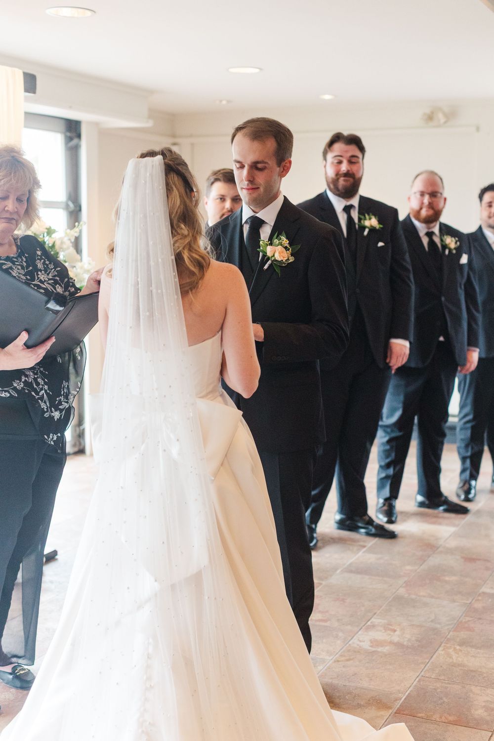 Bride and groom exchange vows at indoor wedding ceremony with groomsmen standing in background.