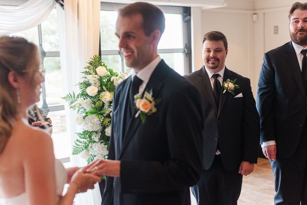 Groom smiling at bride during indoor wedding ceremony with groomsmen and floral arrangement in background.