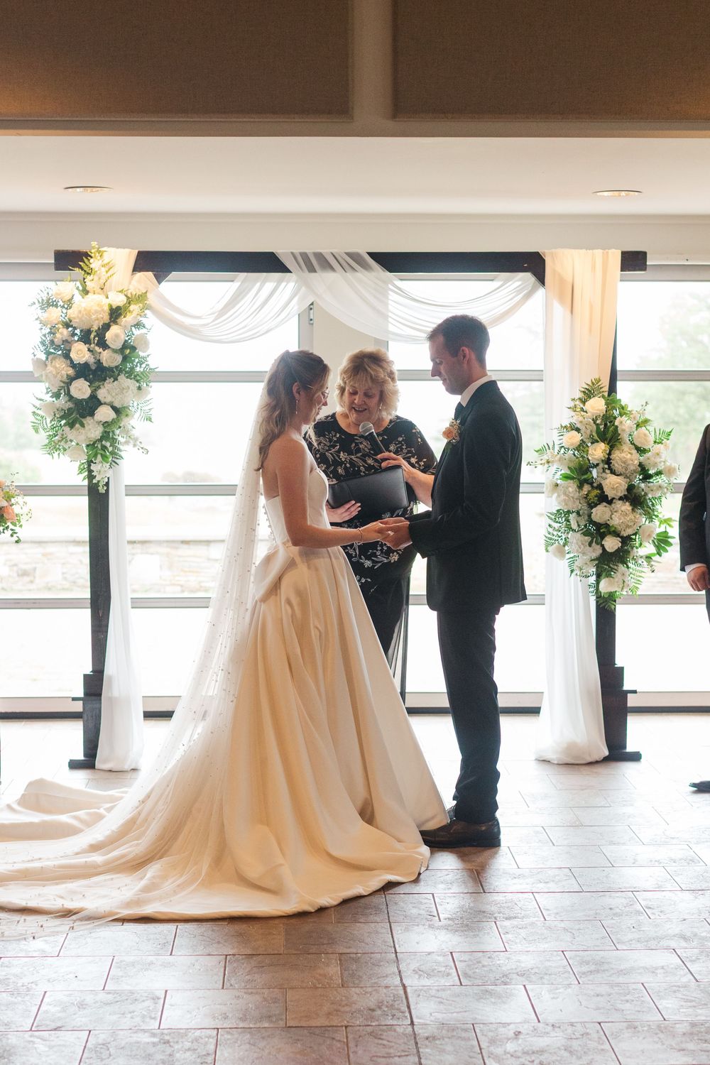 Bride and groom exchange vows at indoor wedding ceremony with white floral arrangements and draped fabric backdrop.