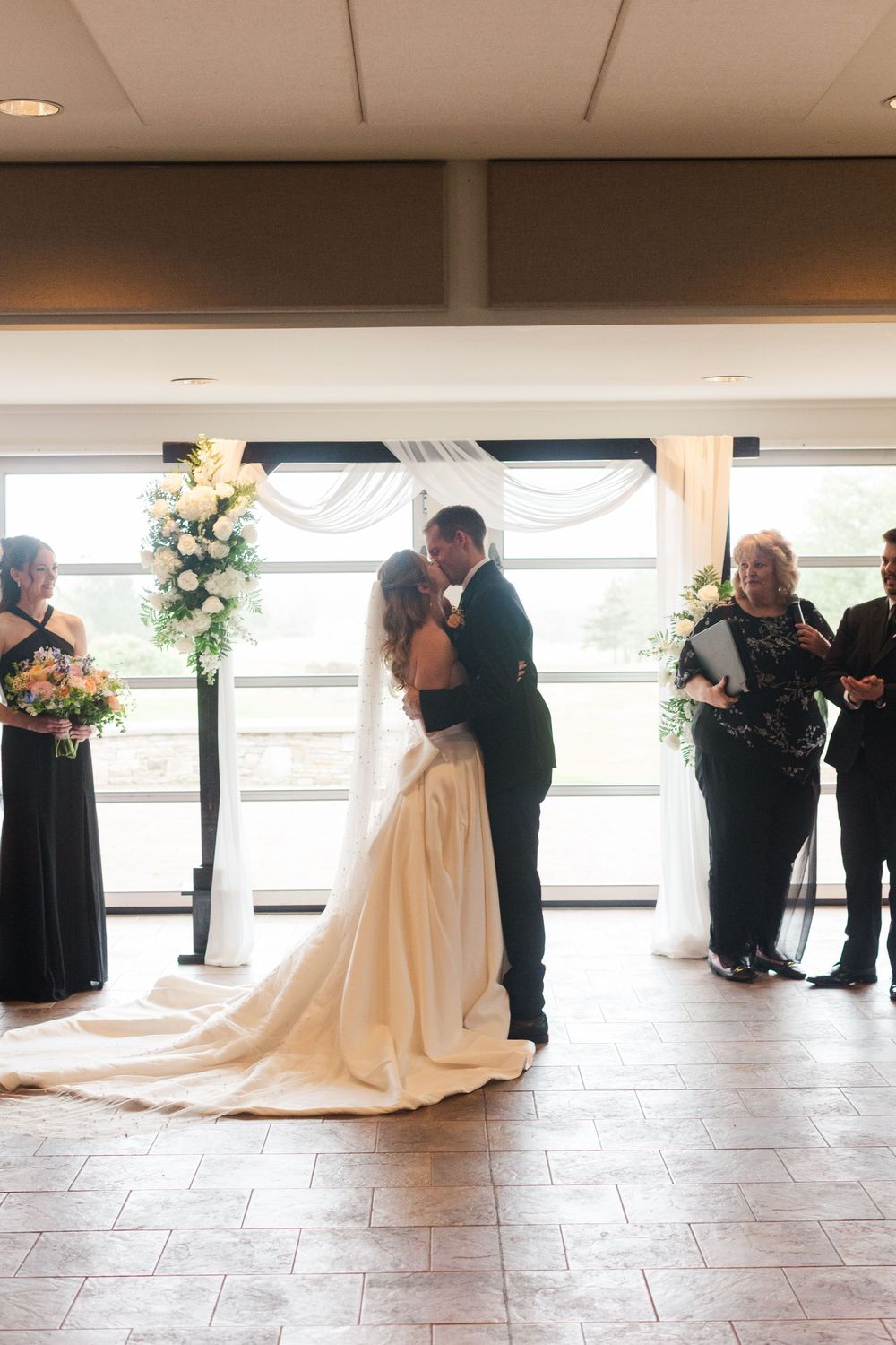 Bride and groom share first kiss at indoor wedding ceremony with floral arch and bridesmaids in black dresses.