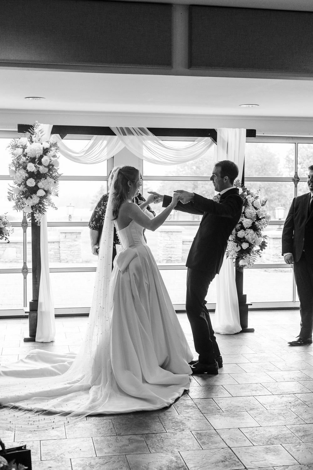 Black and white photo of bride and groom exchanging vows at elegant indoor wedding ceremony with floral arrangements.
