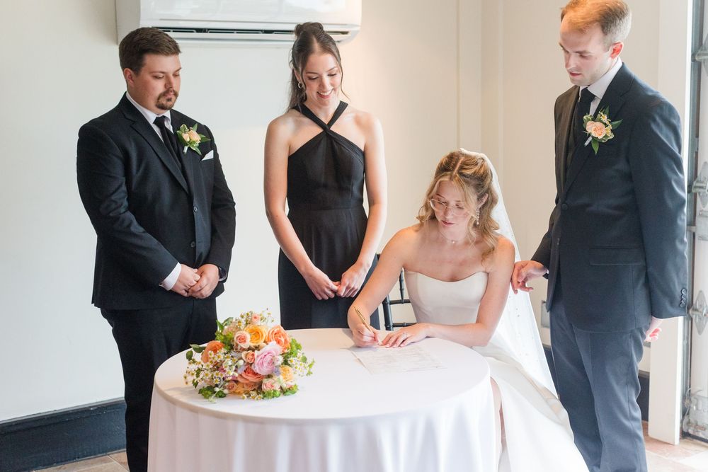 Bride signing wedding documents at a white table surrounded by groom and wedding party in formal attire.