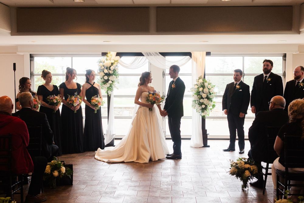 Bride and groom exchange vows at elegant indoor wedding ceremony with bridesmaids in black gowns and white floral arrangements.