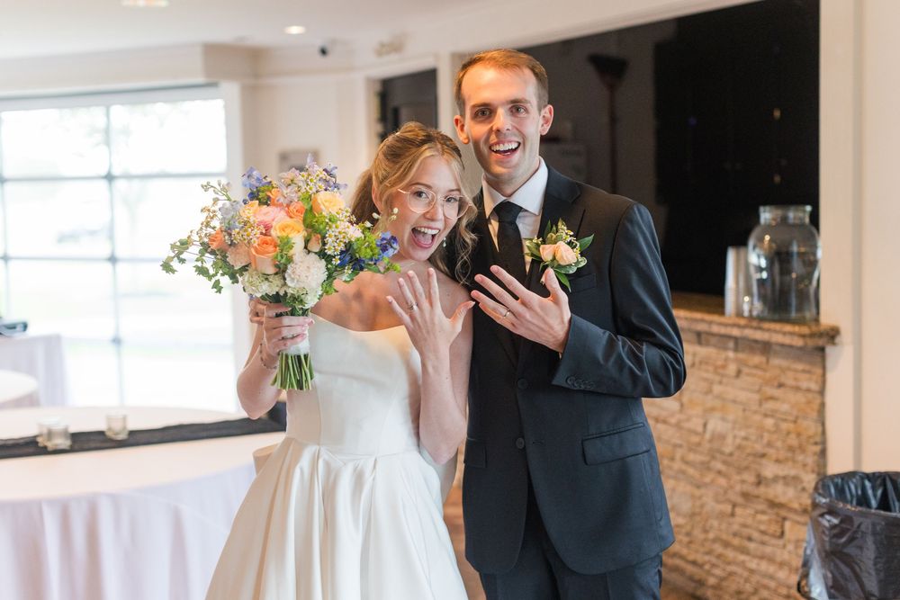 Newlyweds smiling and showing off wedding rings, bride holding colorful bouquet in elegant indoor venue.
