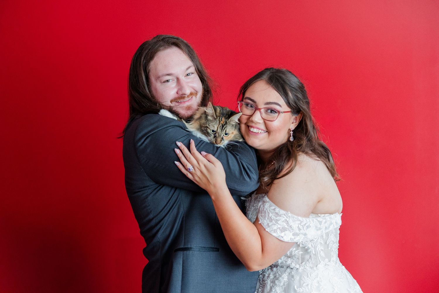 Couple in wedding attire smiling and holding a cat against a vibrant red background.