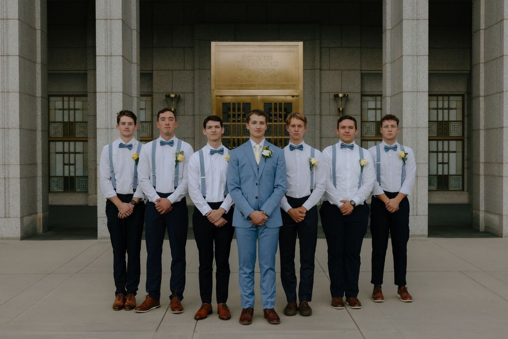 Groom in light blue suit stands with groomsmen in navy pants and suspenders outside a grand building.