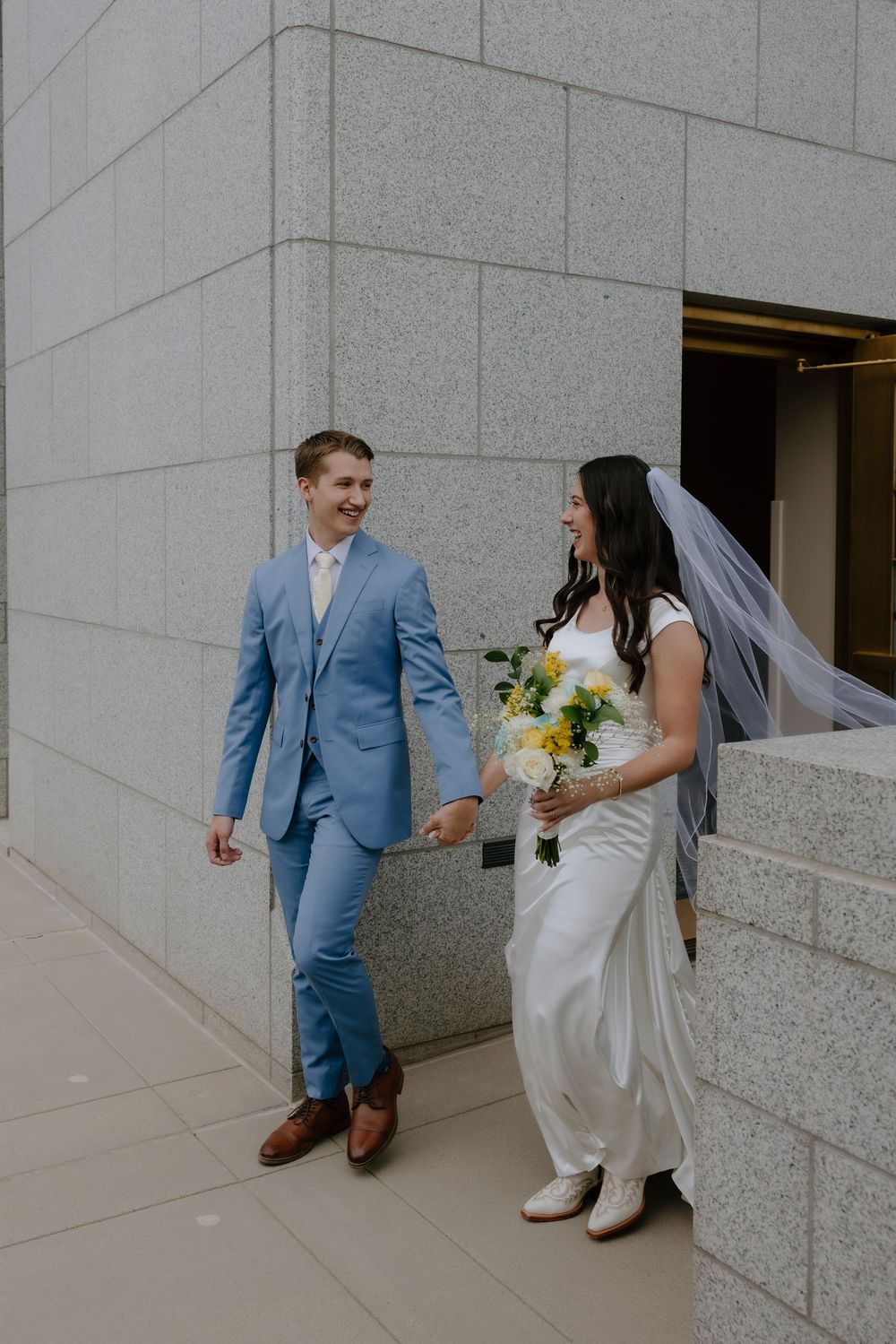Bride in white gown with yellow bouquet and groom in light blue suit hold hands outside a stone building.