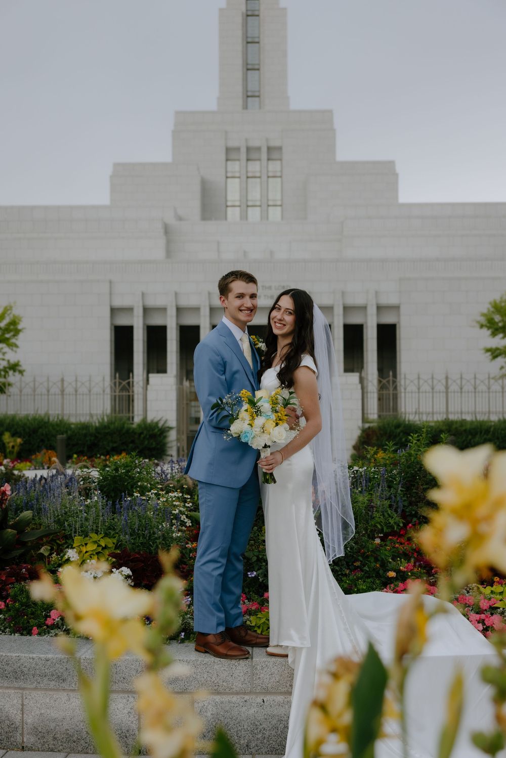 Bride and groom posing in front of an LDS temple surrounded by colorful flower gardens on their wedding day.