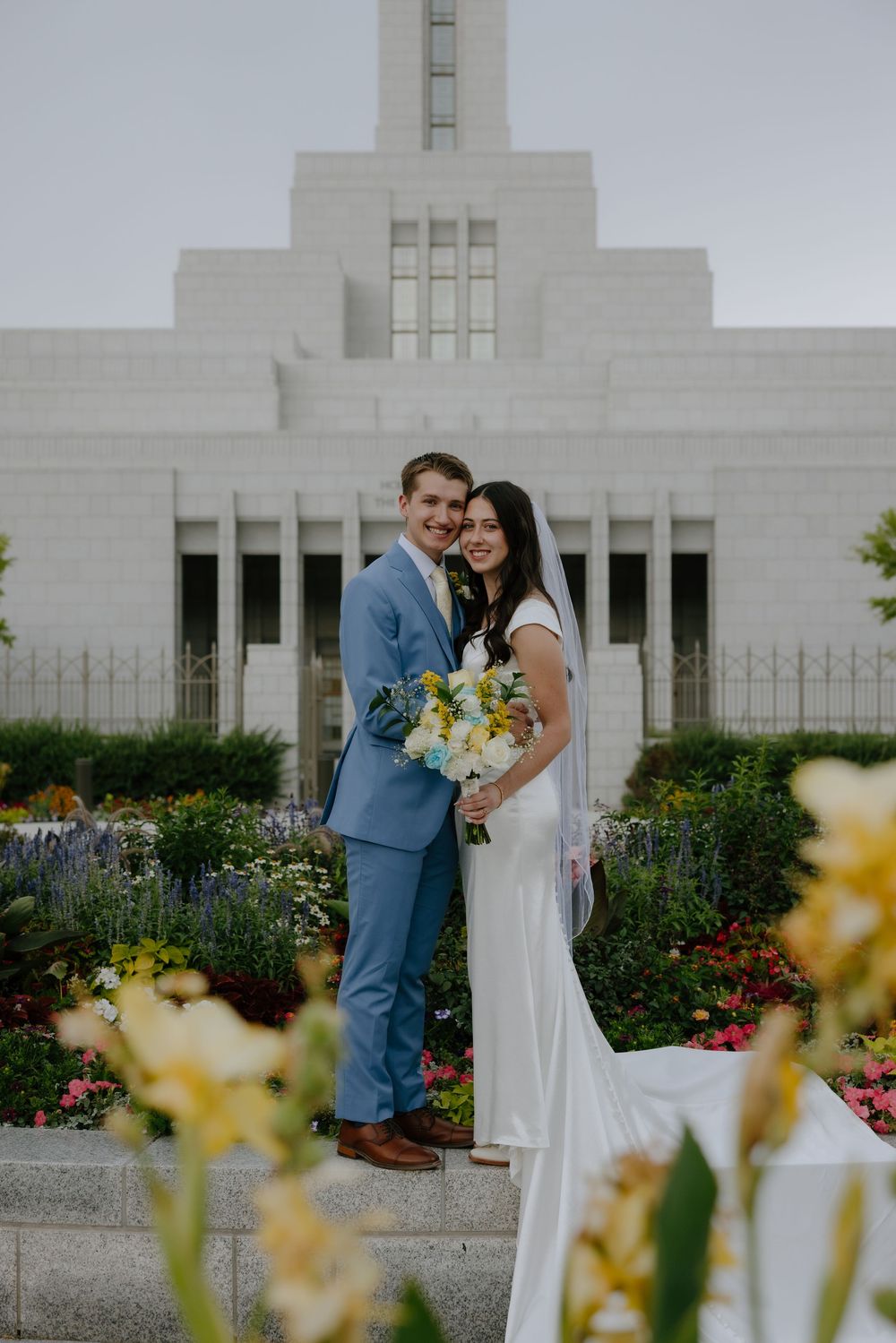 Elegant bride and groom pose together in front of a white LDS temple surrounded by colorful blooming flowers.