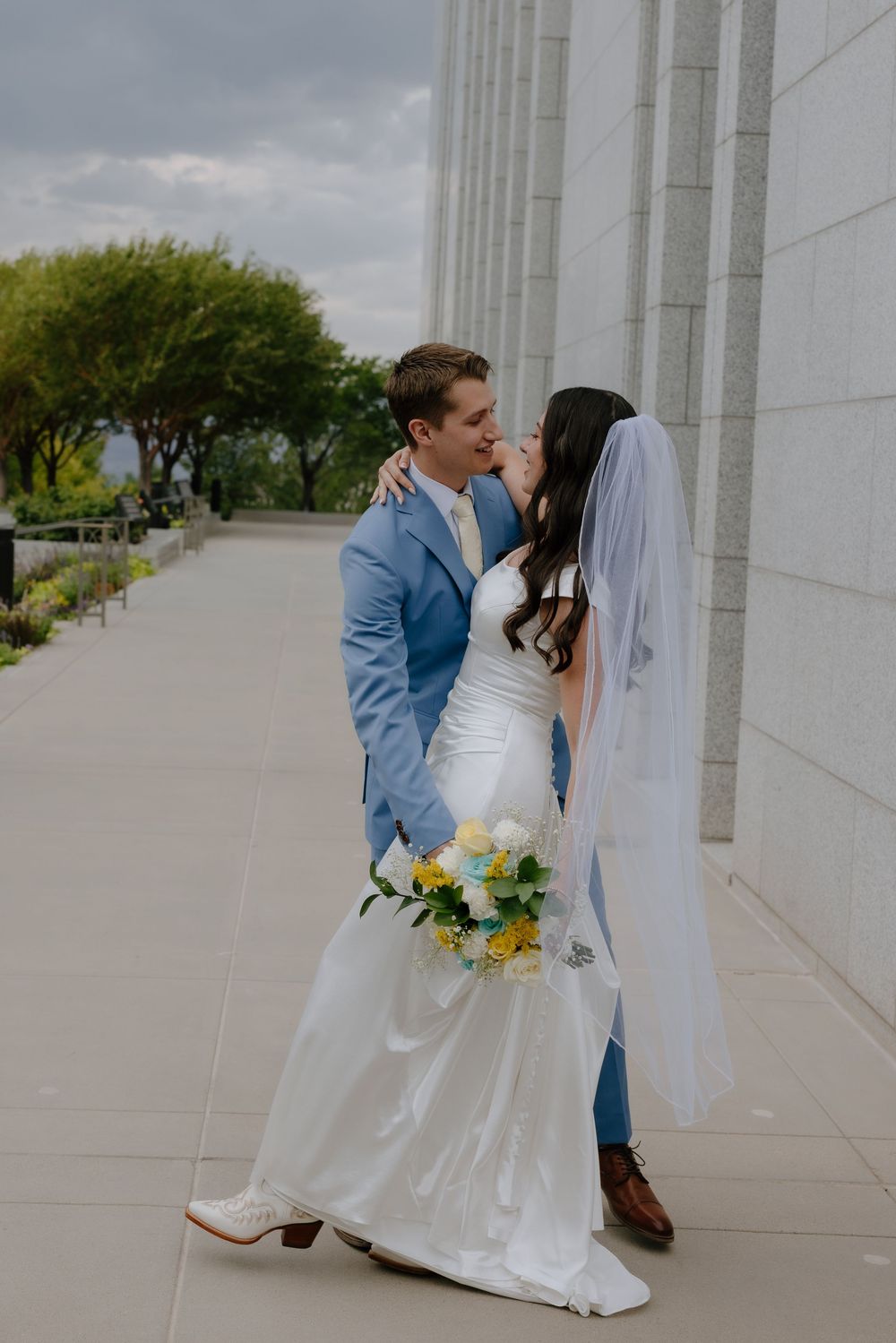 Bride and groom sharing a kiss outdoors near marble columns, bride holding yellow and white bouquet wearing veil.