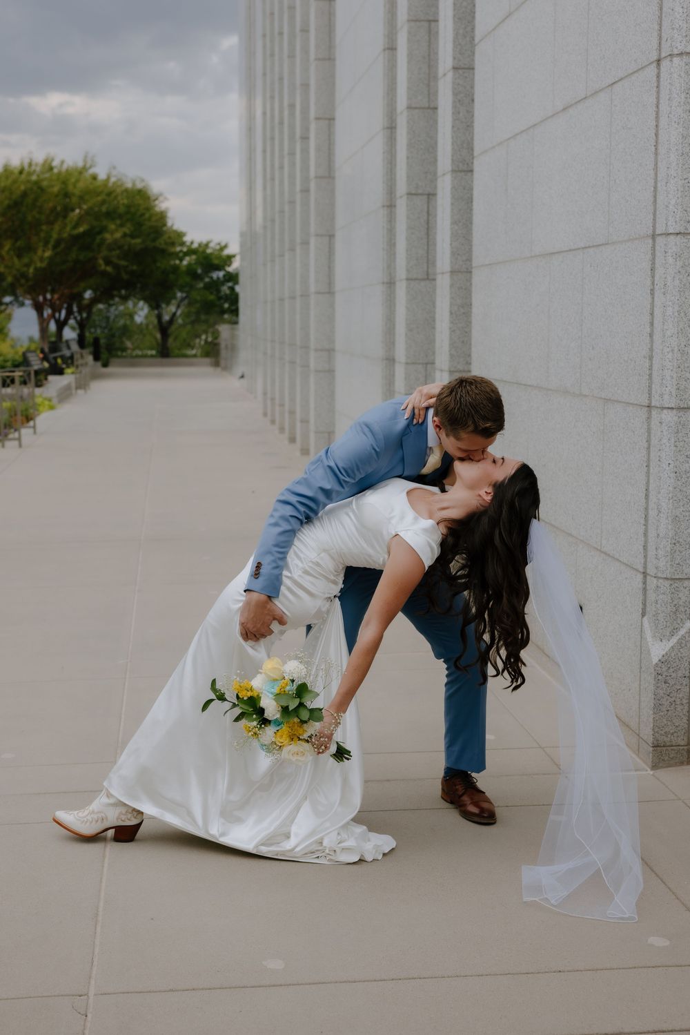 Bride and groom share a romantic dip kiss outside a white stone building, bride holding yellow and white bouquet.