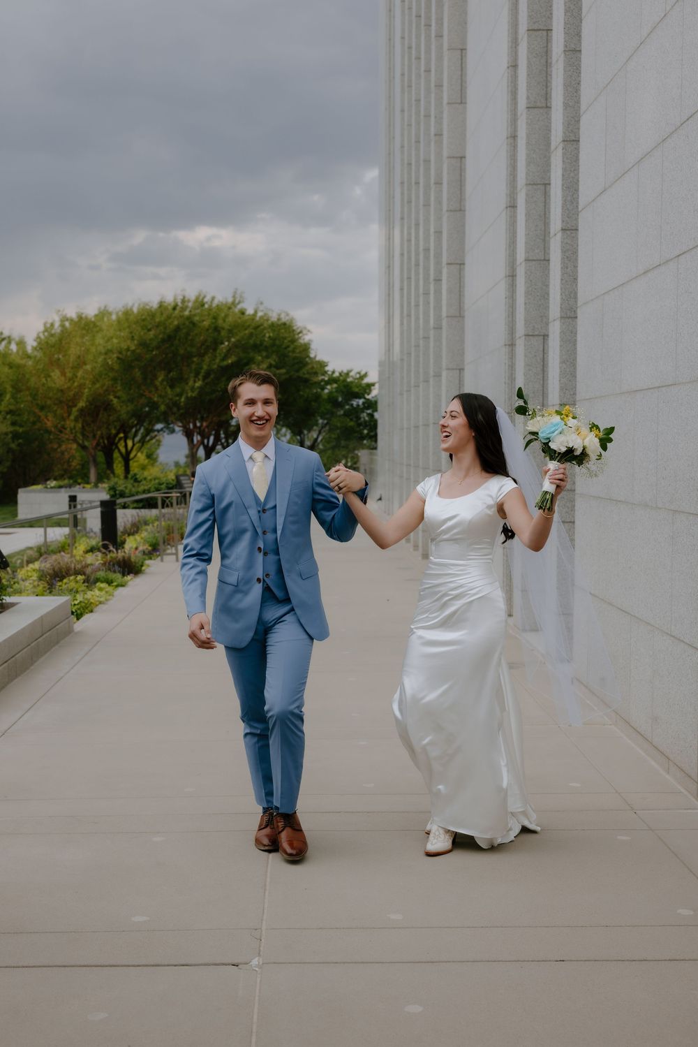 A joyful bride and groom walk hand-in-hand outside a white stone building, she holds a bouquet in a white gown, he wears a blue suit.