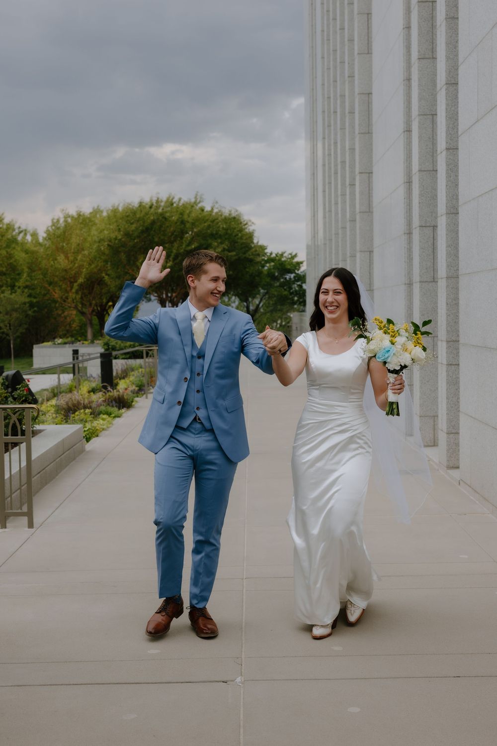 Joyful newlyweds walk together, groom in blue suit waving, bride in white gown holding yellow and blue bouquet.