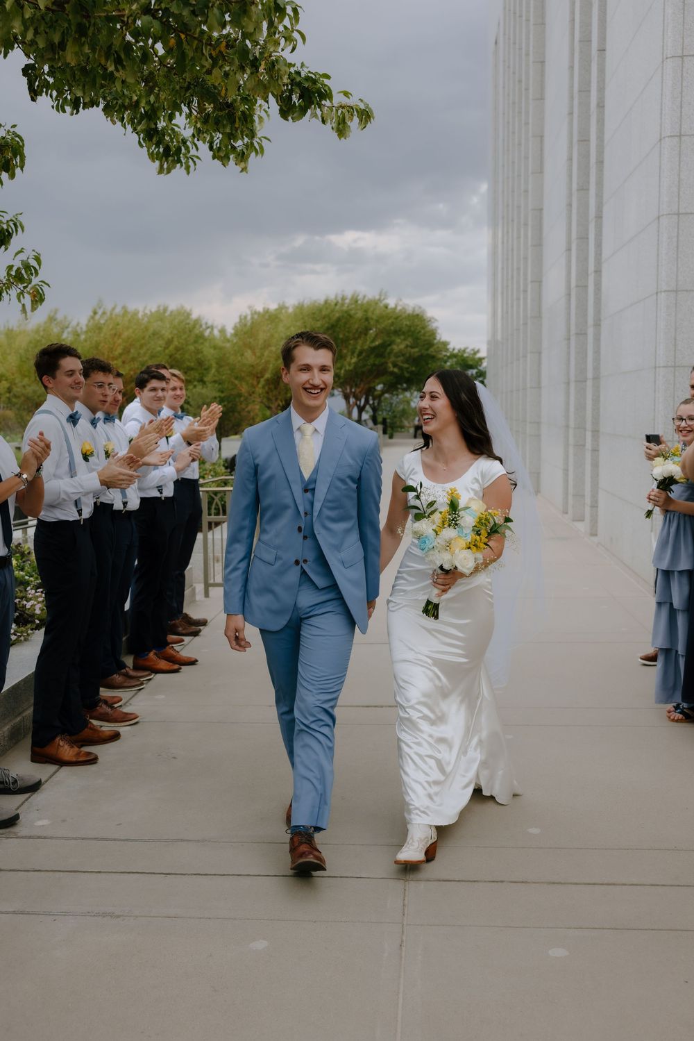 Beaming newlyweds walk hand-in-hand past applauding guests outside a white temple after their wedding ceremony.