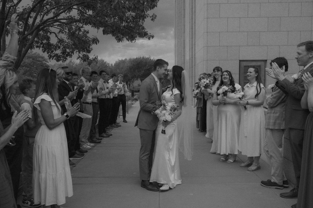 Black and white photo of a newlywed couple kissing as guests line up to celebrate outside a wedding venue.