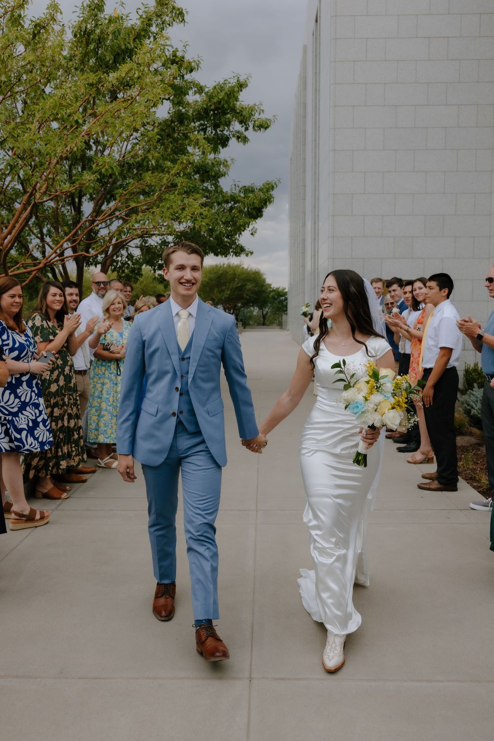 Happy bride and groom hold hands walking past cheering guests outside a white LDS temple on their wedding day.