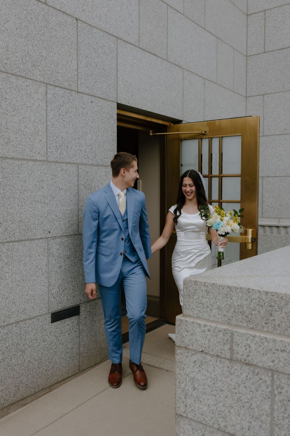 Bride and groom exit golden doors in blue suit and white wedding dress, holding hands and smiling.