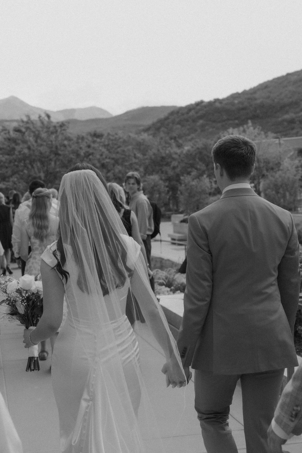 Black and white photo of bride and groom walking hand in hand at outdoor wedding with mountain views.