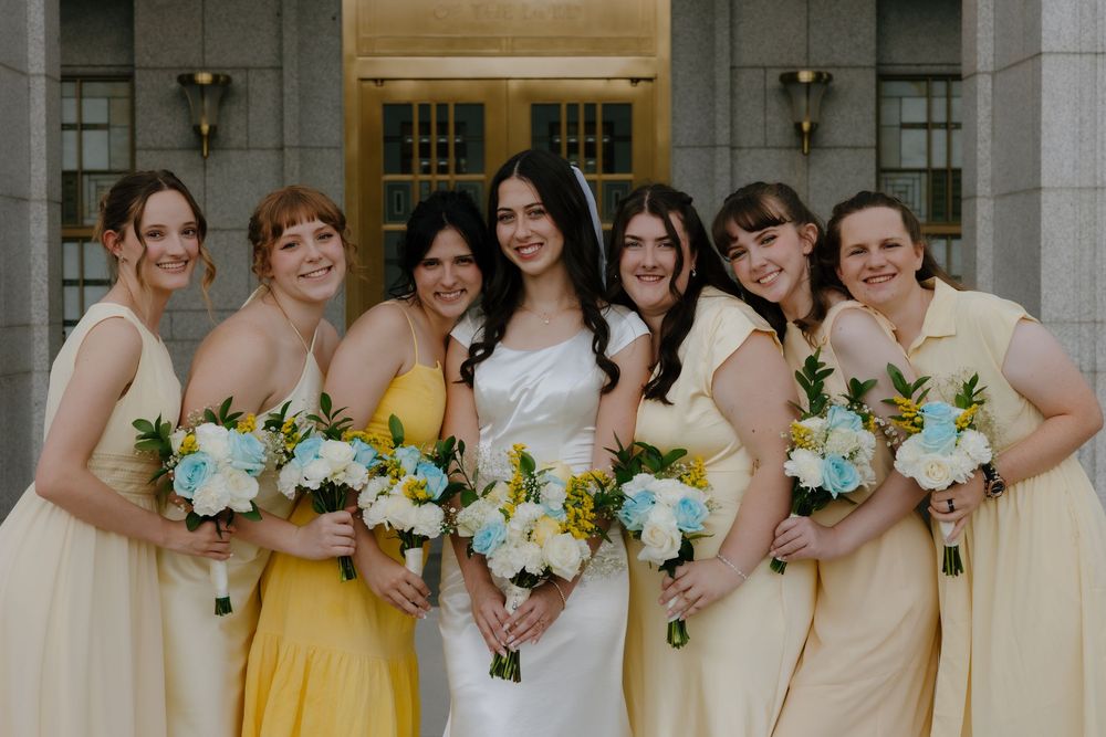 Bride in white gown with six bridesmaids in yellow dresses holding white and blue floral bouquets outdoors.