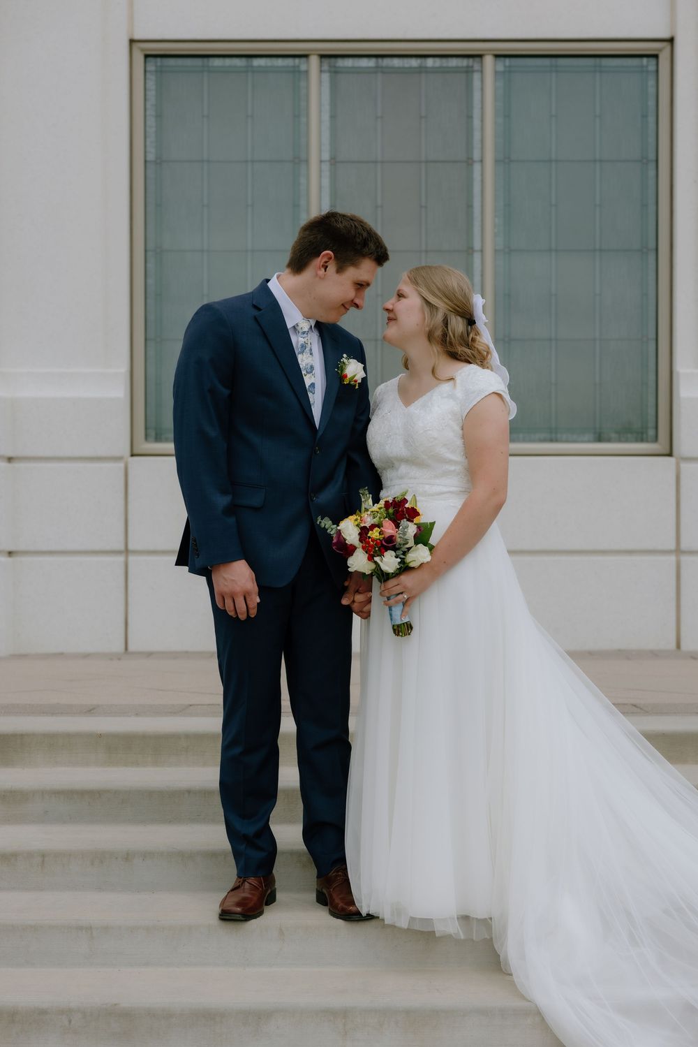 Bride and groom share a loving glance on white marble steps, bride holding colorful bridal bouquet.