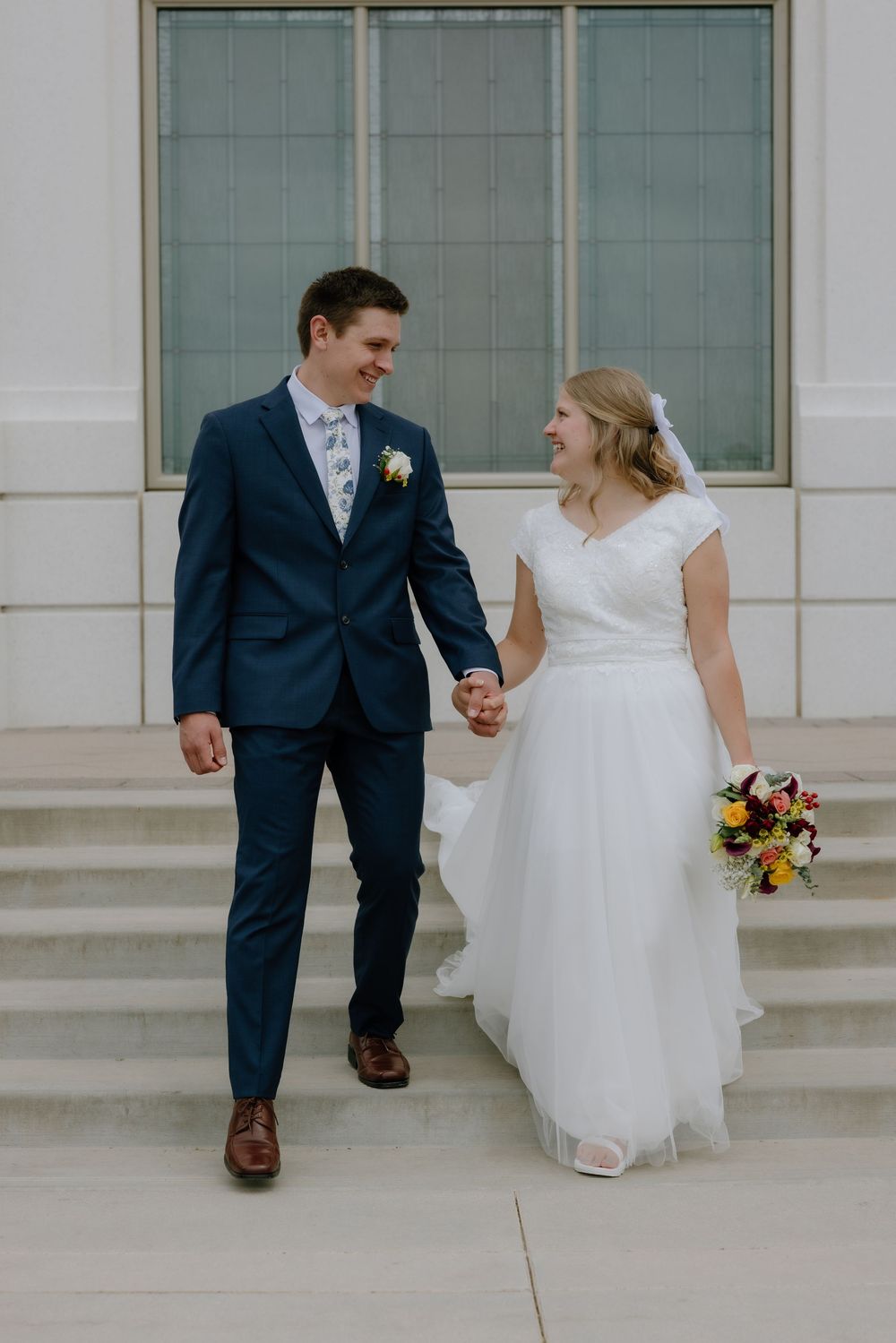 Smiling bride and groom walk hand in hand down steps outside a temple on their wedding day.