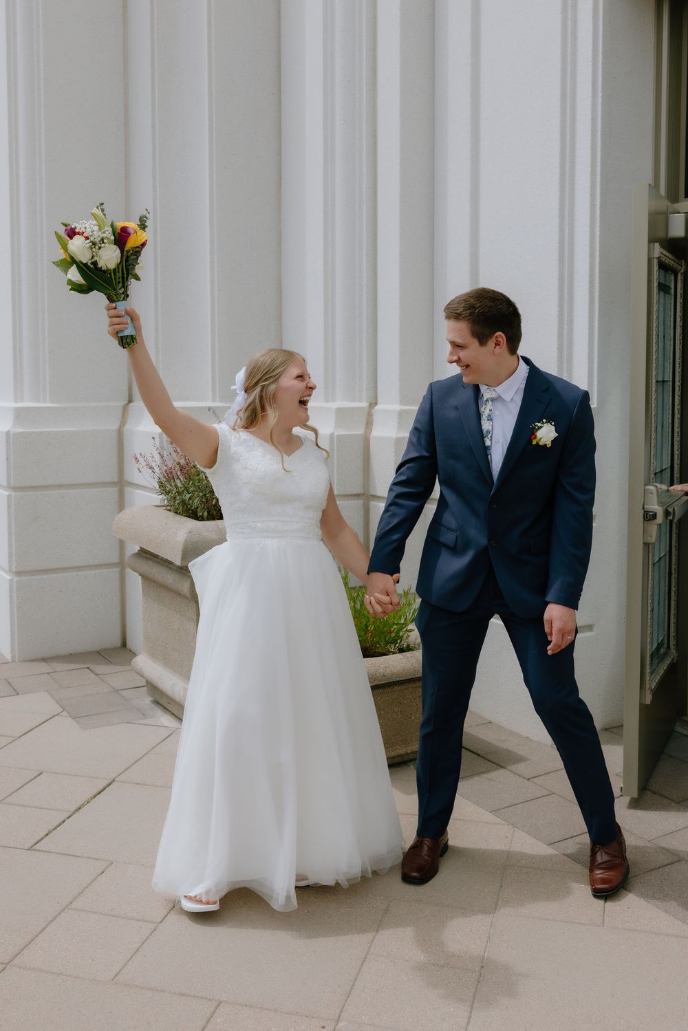 Joyful bride raises colorful bouquet while groom in navy suit holds her hand outside a white building.