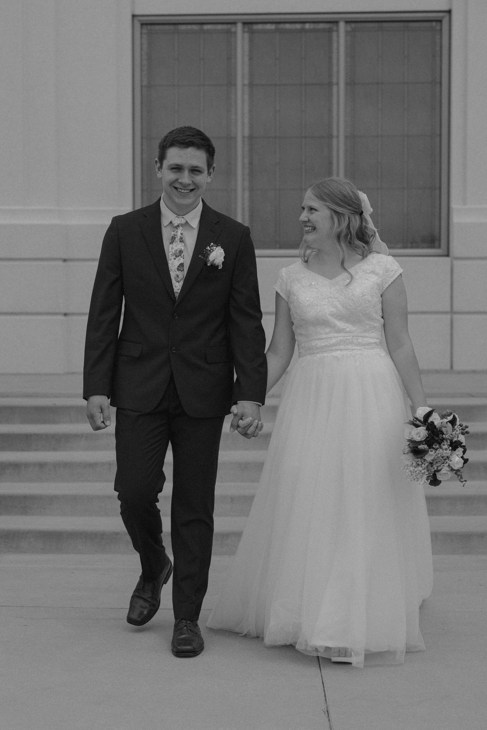 Black and white photo of smiling newlyweds walking hand in hand down temple steps after ceremony.