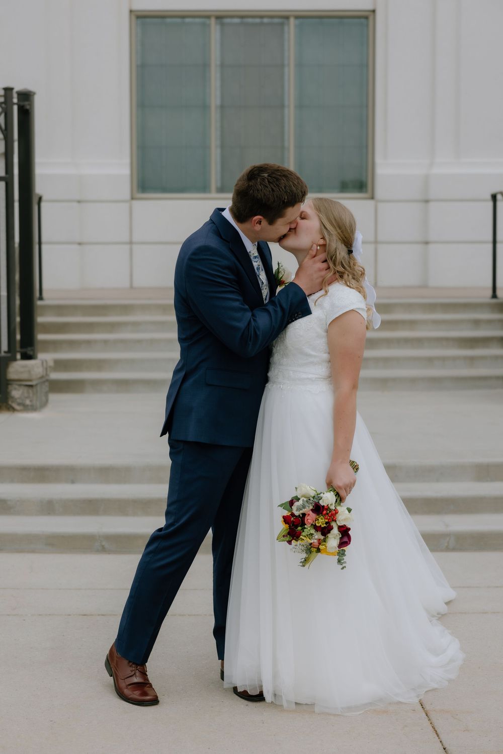 Bride and groom share a passionate kiss outdoors, bride holding vibrant red and yellow bridal bouquet.