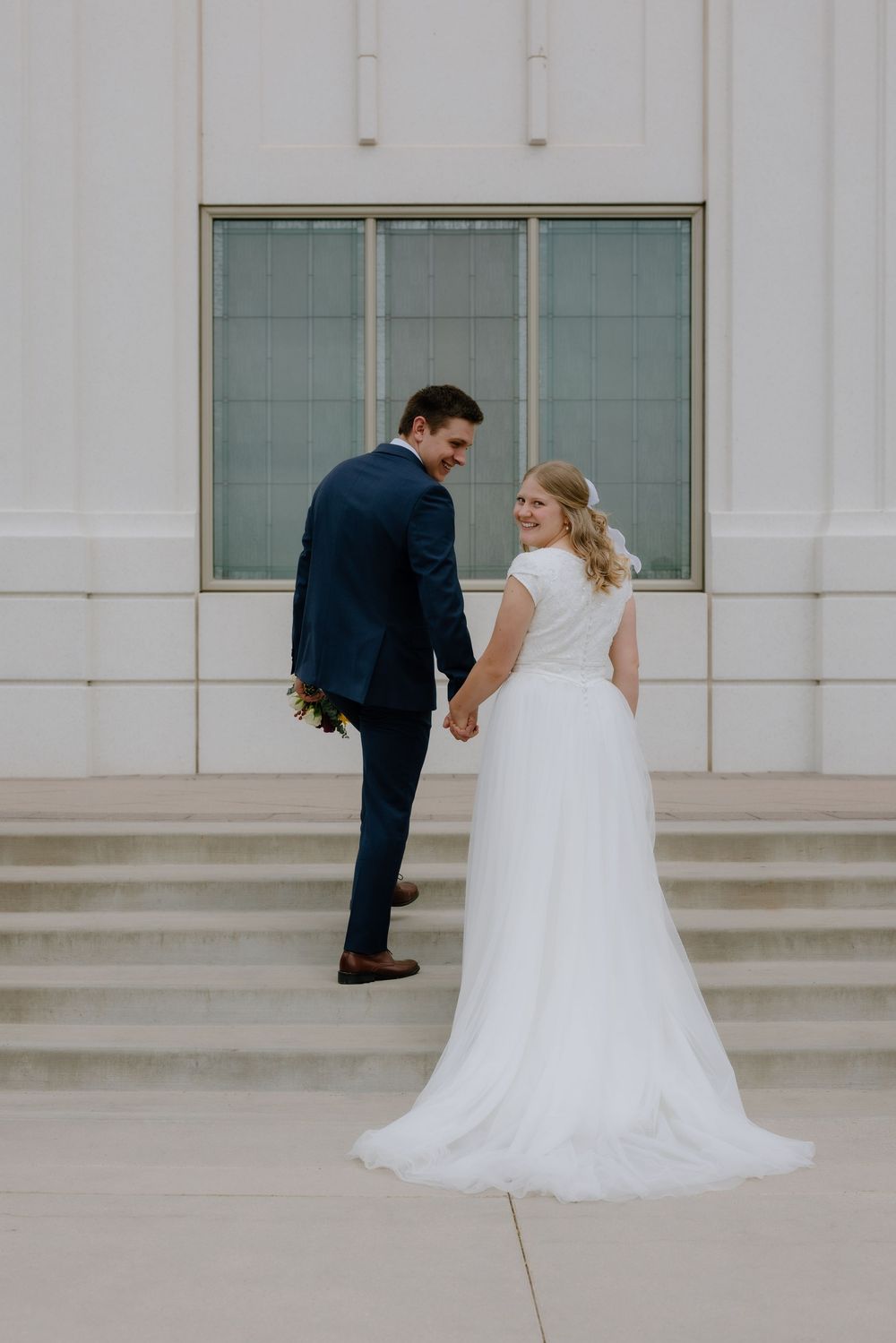 Bride and groom holding hands and smiling while posing on steps outside a grand white temple.