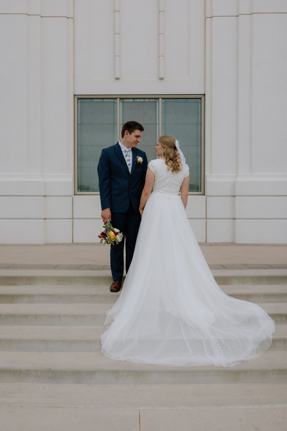 Bride and groom pose on temple steps, showcasing her flowing white gown train and his navy blue suit.