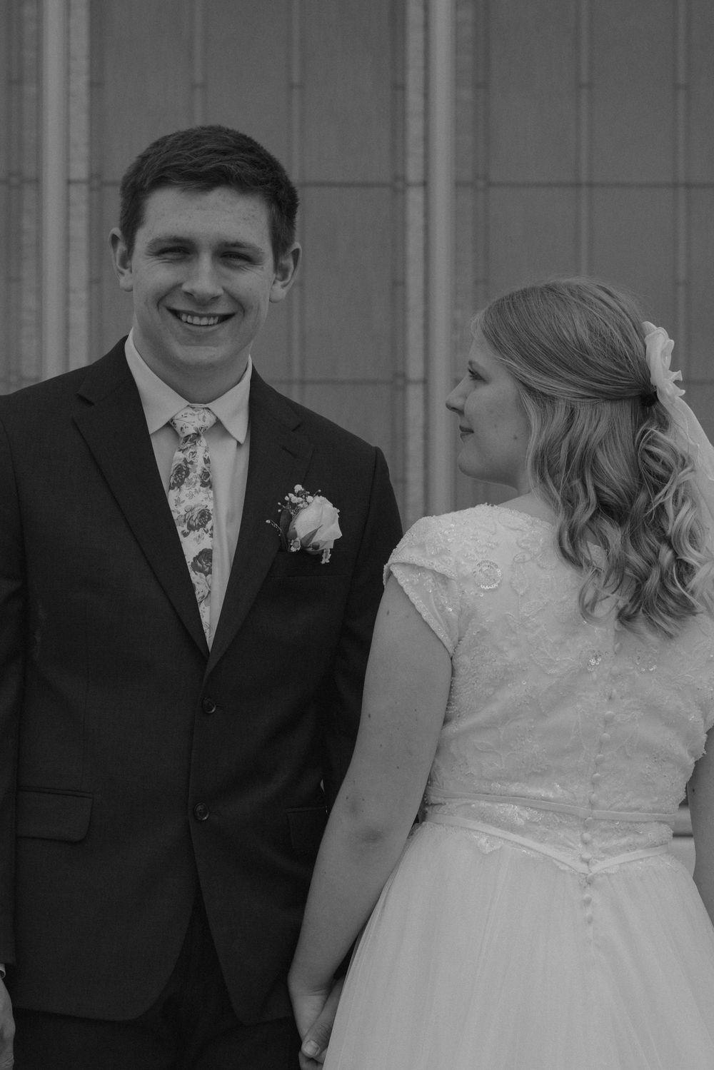 Black and white portrait of smiling groom in suit and bride in lace wedding dress holding hands.