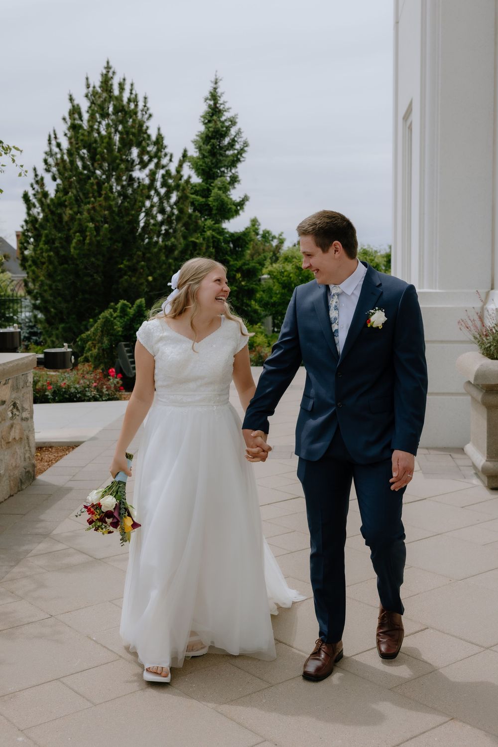 Smiling bride and groom walk hand in hand on a temple pathway with lush green trees behind them.