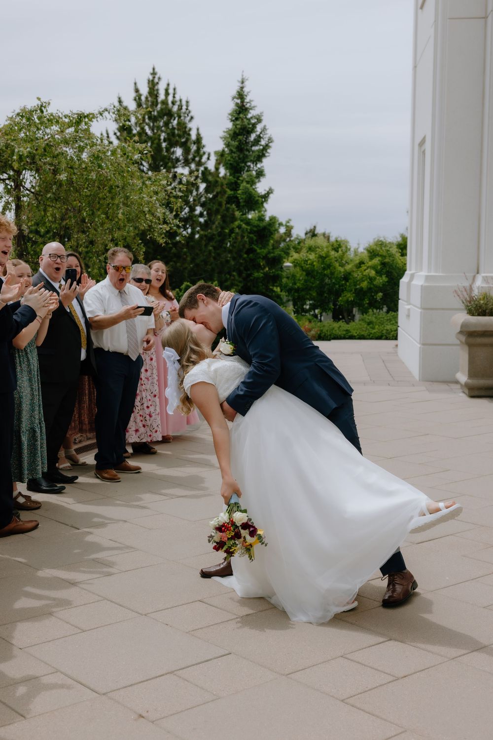 Groom dips bride in a romantic kiss as wedding guests applaud on a temple courtyard walkway.