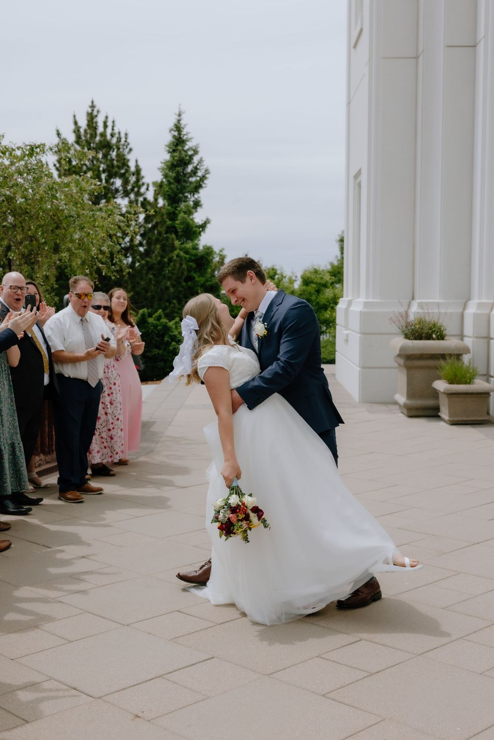 Groom dips bride for a kiss while guests celebrate on a bright outdoor temple courtyard.