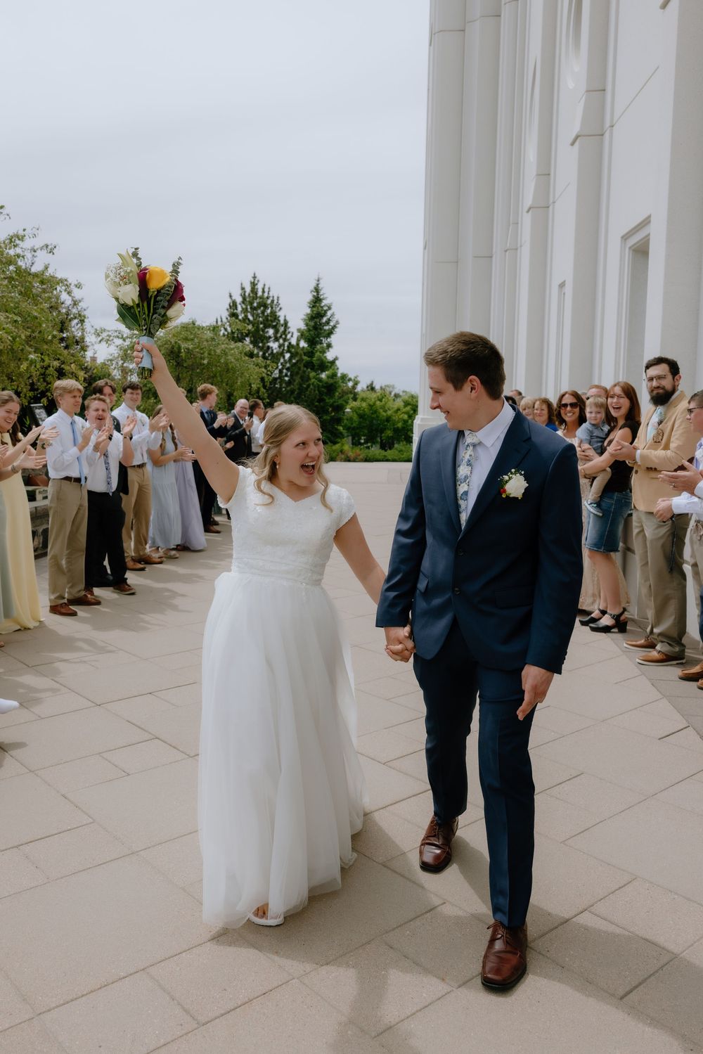 Joyful bride raises bouquet while walking with groom through cheering guests outside white temple.