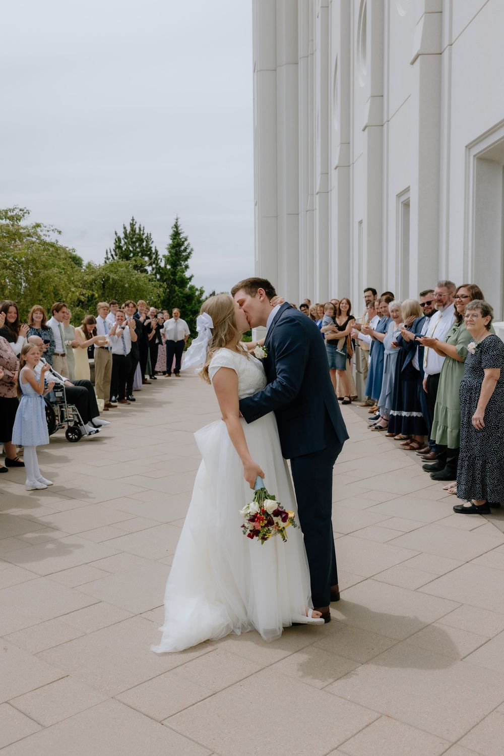 Newlyweds sharing a romantic kiss outside a white temple surrounded by cheering wedding guests.