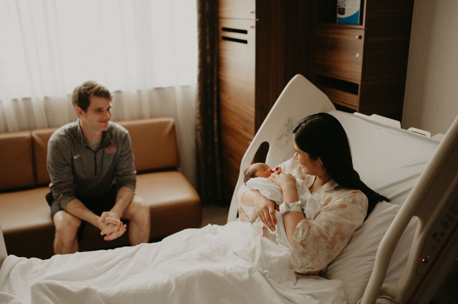 New mother resting in a hospital bed holding her newborn baby while a smiling father sits nearby on a couch.