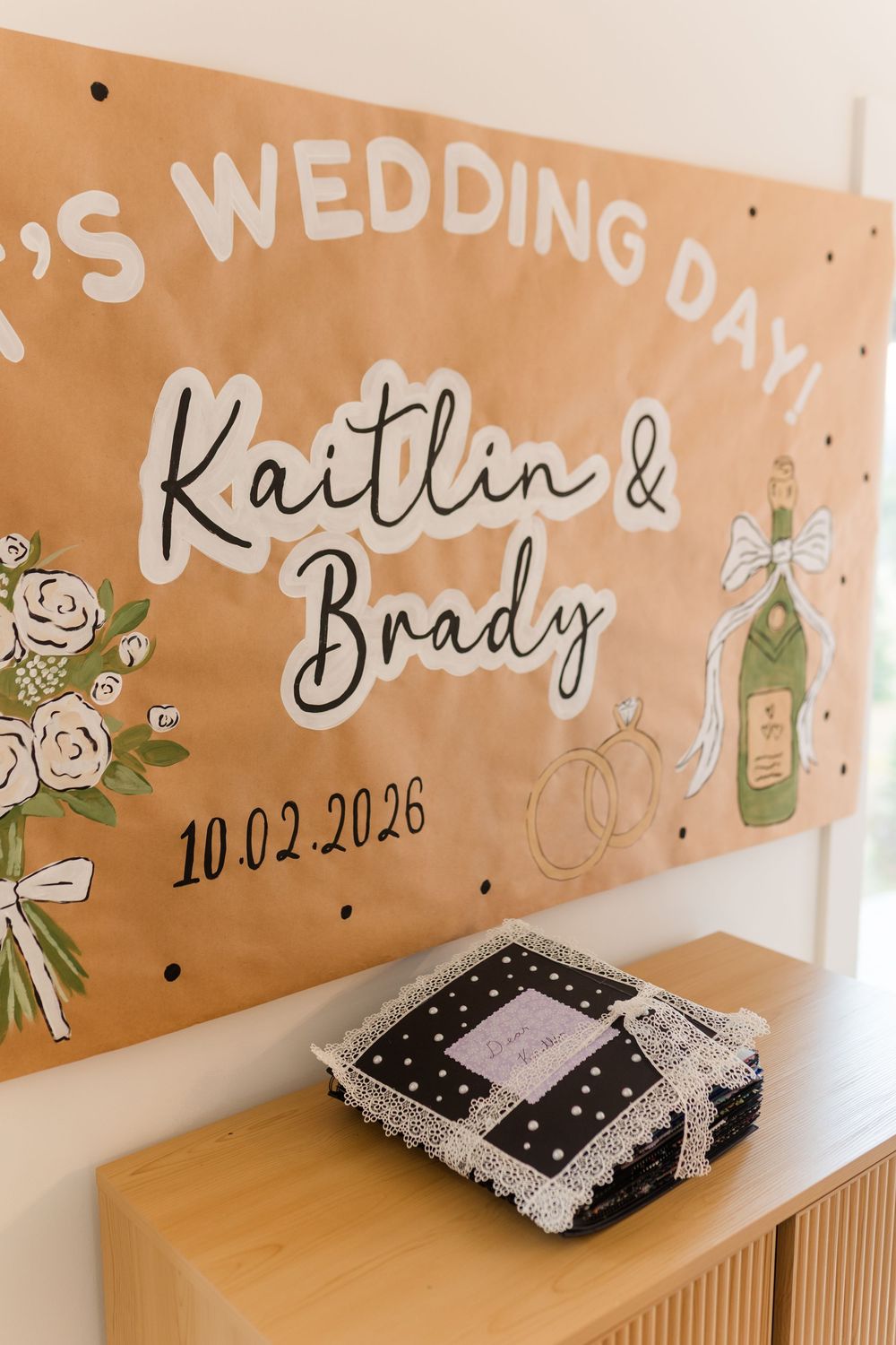 Hand-painted kraft paper wedding banner for Kaitlin & Brady dated 10.02.2026 with florals, rings and champagne above a wooden console.