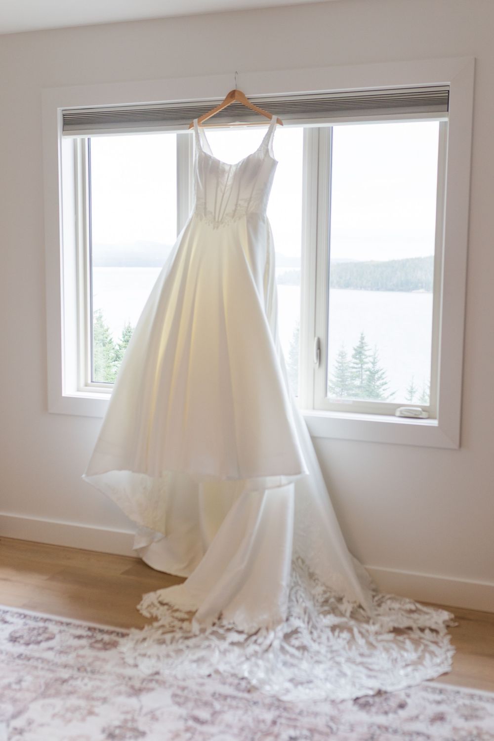 White satin wedding dress with lace-trimmed train hanging from a window overlooking a lake and evergreen trees.