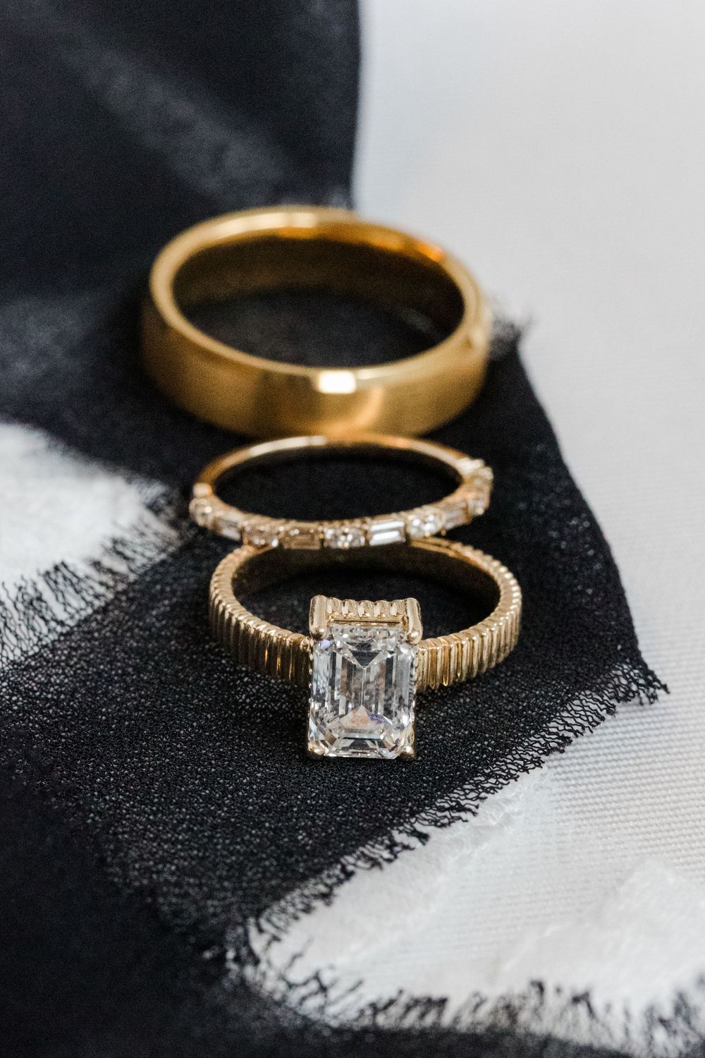 Three gold wedding rings with an emerald-cut diamond engagement ring, a baguette band, and a plain band resting on black and white fabric.