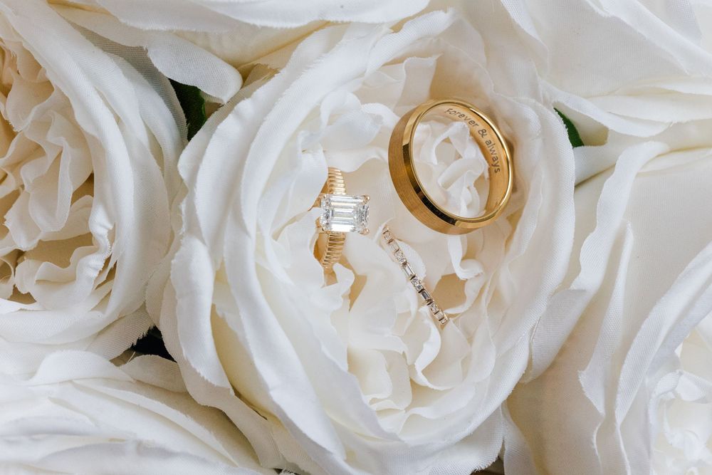 Diamond engagement ring and gold wedding band resting on soft white garden roses, a romantic wedding detail photo.