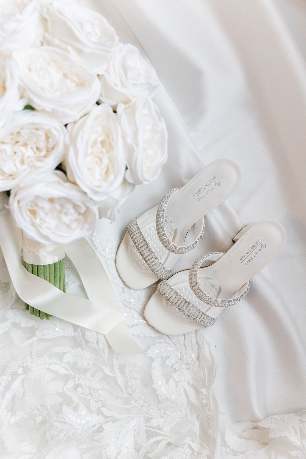 White bridal bouquet of roses with ivory ribbon beside embellished open-toe wedding sandals on a lace wedding dress.